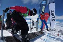 Snowboarder an einem Startplatz im Schnee, mit Helm und Startnummern, neben Fahnen vor verschneiten Bergen.