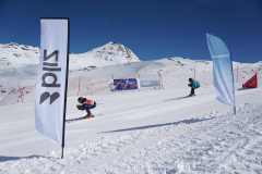 Zwei Skifahrer auf einer präparierten Abfahrt zwischen Werbefahnen vor verschneiten Bergen.