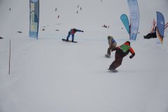 Snowboarder bei einem Rennen auf einer verschneiten Piste mit Ziellinie und Werbefahnen.