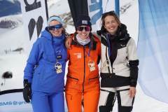 Drei Wintersportlerinnen in Skianzügen stehen mit Medaillen vor verschneiter Bergkulisse.