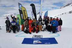 Gruppe von Wintersportlern posiert auf einem verschneiten Berggipfel vor Fahnen und einem Schneesport-Team-Banner.