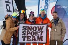 Gruppe von fünf Personen in Winterkleidung hält ein „SNOW SPORT TEAM“-Banner vor verschneiter Bergkulisse.