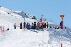 Skifahrer stehen am Start eines verschneiten Hanges unter blauem Himmel.
