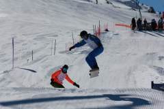 Snowboarder springt über einen anderen Fahrer auf einer präparierten Piste im Schnee.