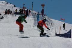 Zwei Snowboarder springen parallel über eine präparierte Piste im Skigebiet.
