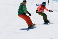 Zwei Snowboarder fahren nebeneinander einen verschneiten Hang hinunter.