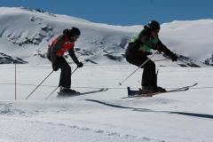 Zwei Skifahrer springen gemeinsam über eine kleine Schneekante auf einer Piste in den Bergen.