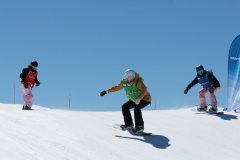 Drei Snowboarder fahren eine verschneite Piste bei klarem blauem Himmel hinunter.