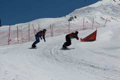 Zwei Snowboarder fahren eine präparierte Schneepiste mit Kurve und Absperrnetz entlang.