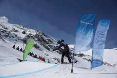 Snowboarder auf einer verschneiten Piste zwischen zwei blauen und einer grünen Fahne vor Bergen.