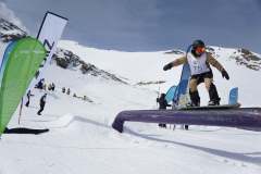Snowboarder mit Startnummer 75 auf einer Rail, daneben Fahnen und weitere Personen im Schnee.