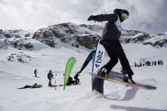 Snowboarder auf einem Rail im Schnee vor Bergkulisse, weitere Personen und Fahnen im Hintergrund.
