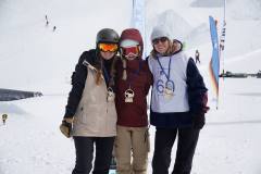 Drei Wintersportlerinnen mit Medaillen stehen nebeneinander im Schnee bei einem Skigebiet.