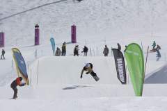 Snowboarder springt über eine Schnee­rampe in einem alpinen Park, umgeben von Fahnen und Zuschauern.