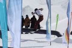 Snowboarder beim Balancieren auf einem Rail im Snowpark, umgeben von Fahnen und Zuschauern.
