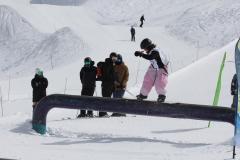 Skifahrerin in pinker Hose auf einer dunklen Rail im Schnee, daneben mehrere Ski- und Snowboardfahrer.