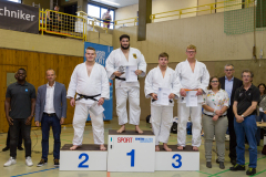 Siegerehrung in einer Sporthalle: fünf Männer auf einem Podest, andere Personen daneben, mehrere halten Urkunden.