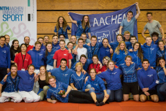 Große Gruppe in blauen Sportshirts vor Bannern, einige stehen, andere sitzen oder knien.