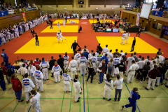 Mehrere Judo-Wettkampfflächen in einer Sporthalle, viele Kämpfer, Trainer und Zuschauer, Kampfrichter und Tische.