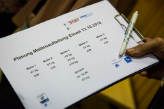 Hand hält Klemmbrett mit Papier „Planung Mattenaufteilung Einzel 13.10.2018“ und Stift.