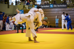 Zwei Judoka im weißen Anzug ringen auf gelber Matte in einer Sporthalle, Zuschauer im Hintergrund.