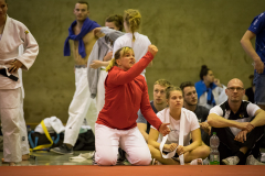 Mehrere Personen in Trainingskleidung in einer Halle; eine Frau im roten Oberteil kniet vorn, andere sitzen und stehen.