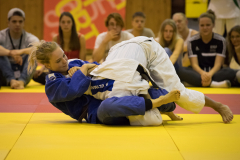 Zwei Judoka in blauen und weißen Anzügen ringen auf gelber Matte, Zuschauer sitzen im Hintergrund.