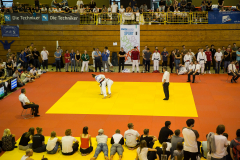 Judo-Kampf auf gelber Matte in Halle; zwei Kämpfer, Kampfrichter und Zuschauer ringsum.