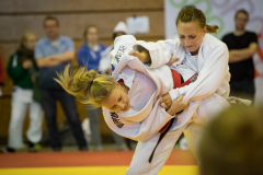 Zwei Judoka im weißen Anzug beim Griffkampf, eine hebt die andere in einer Halle.