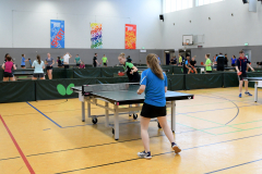 Mehrere Personen spielen Tischtennis in einer Sporthalle; im Vordergrund steht eine Frau an einer Tischtennisplatte.