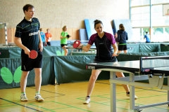 Zwei Personen spielen Tischtennis in einer Halle, mit Schlägern an einem Tisch; weitere Spieler stehen im Hintergrund.