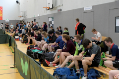 Zuschauer auf einer Bank entlang einer Hallenwand, mit Sporttaschen, Basketballkörben und einer Tür mit Schild „Geräteraum 2“.