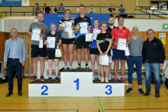 Gruppenfoto bei einer Siegerehrung in einer Turnhalle, Personen auf Podestplätzen mit Urkunden und Tüten.