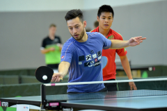 Mann in blauem Shirt spielt Tischtennis; Ball und Schläger über der Platte, zwei weitere Spieler im Hintergrund.