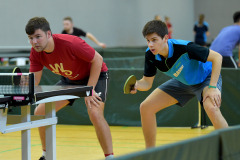 Zwei Tischtennisspieler an einer Tischtennisplatte in einer Halle, einer rot, einer blau-schwarz gekleidet.