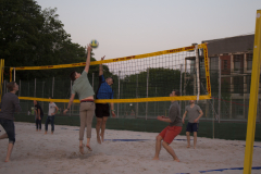 Mehrere Personen spielen Beachvolleyball auf einem Sandplatz; ein Ball fliegt über das Netz.