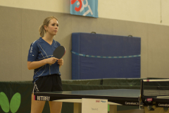 Frau mit Tischtennisschläger neben einer Tischtennisplatte in einer Halle.