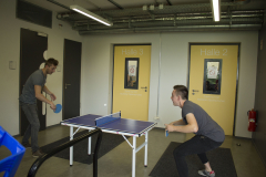 Zwei Männer spielen Tischtennis in einem Flur vor gelben Türen „Halle 2“ und „Halle 3“.