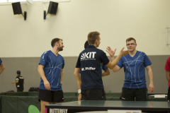 Drei Männer in blauen Sportshirts stehen an einer Tischtennisplatte; einer trägt ein dunkelblaues Shirt mit „KIT“.