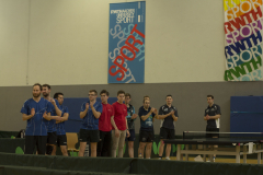 Mehrere Personen stehen und klatschen in einer Halle; Tischtennisplatte rechts, Banner an der Wand.