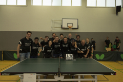 Gruppe von Personen hinter einer Tischtennisplatte in einer Halle; Anzeigetafel zeigt 6:0.