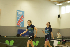 Zwei Frauen beim Tischtennis in einer Halle, eine mit Schläger und Ball, im Hintergrund ein Spieler und ein Banner.