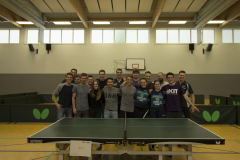 Gruppe von 13 Personen in einer Sporthalle hinter einer Tischtennisplatte, Basketballkorb an der Wand.