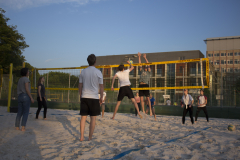 Menschen spielen Volleyball auf einem Sandplatz; ein Spieler springt am Netz, Ball rechts unten.