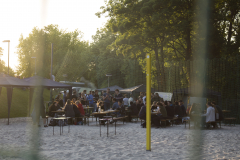 Menschen sitzen an Biertischen im Sand, daneben Sonnenschirme, ein gelber Pfosten und Bäume im Hintergrund.