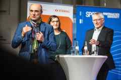 Drei Personen auf einer Podiumsdiskussion, ein Mann spricht ins Mikrofon.