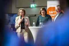 Drei Personen auf einer Bühnenveranstaltung, eine Frau spricht ins Mikrofon.