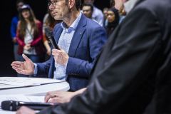 Ein Mann im blauen Anzug spricht mit Mikrofon an einem Podium, während im Hintergrund weitere Personen stehen.