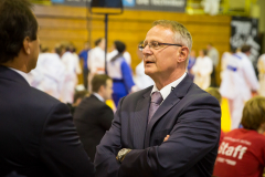 Zwei Männer im Anzug im Gespräch in einer Sporthalle; im Hintergrund Personen in weißen und blauen Anzügen.