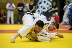 Zwei Judoka in weißen Anzügen ringen auf einer gelben Matte, Zuschauer stehen im Hintergrund.